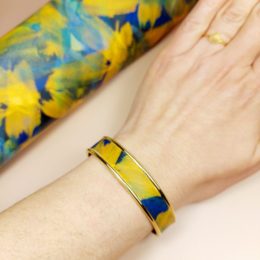 bracelet jonc manchette doré à l'or fin 24k au poignet d'une femme avec lanière cuir bicolore jaune et bleu